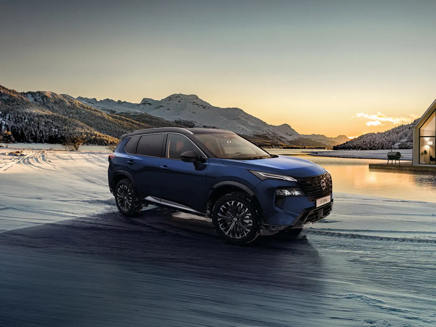 Nissan X-TRAIL SUV 7 Places bleu roulant dans un paysage enneigé près d'un lac avec des montagnes au coucher du soleil.