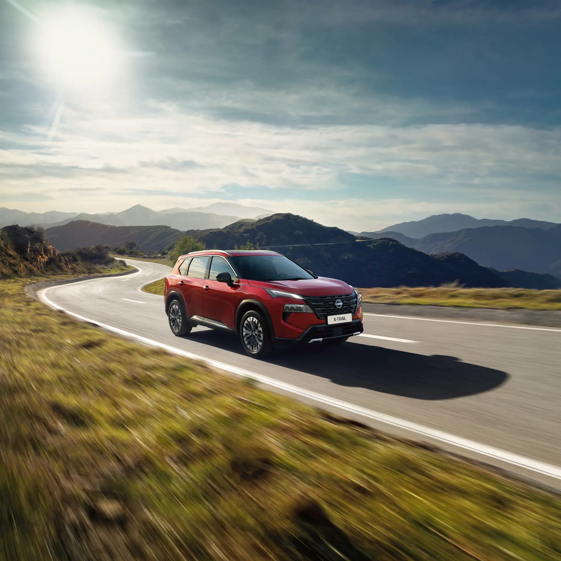 Nissan X-TRAIL SUV 7 Places rouge roulant le long d'une route de montagne pittoresque sous un ciel lumineux.