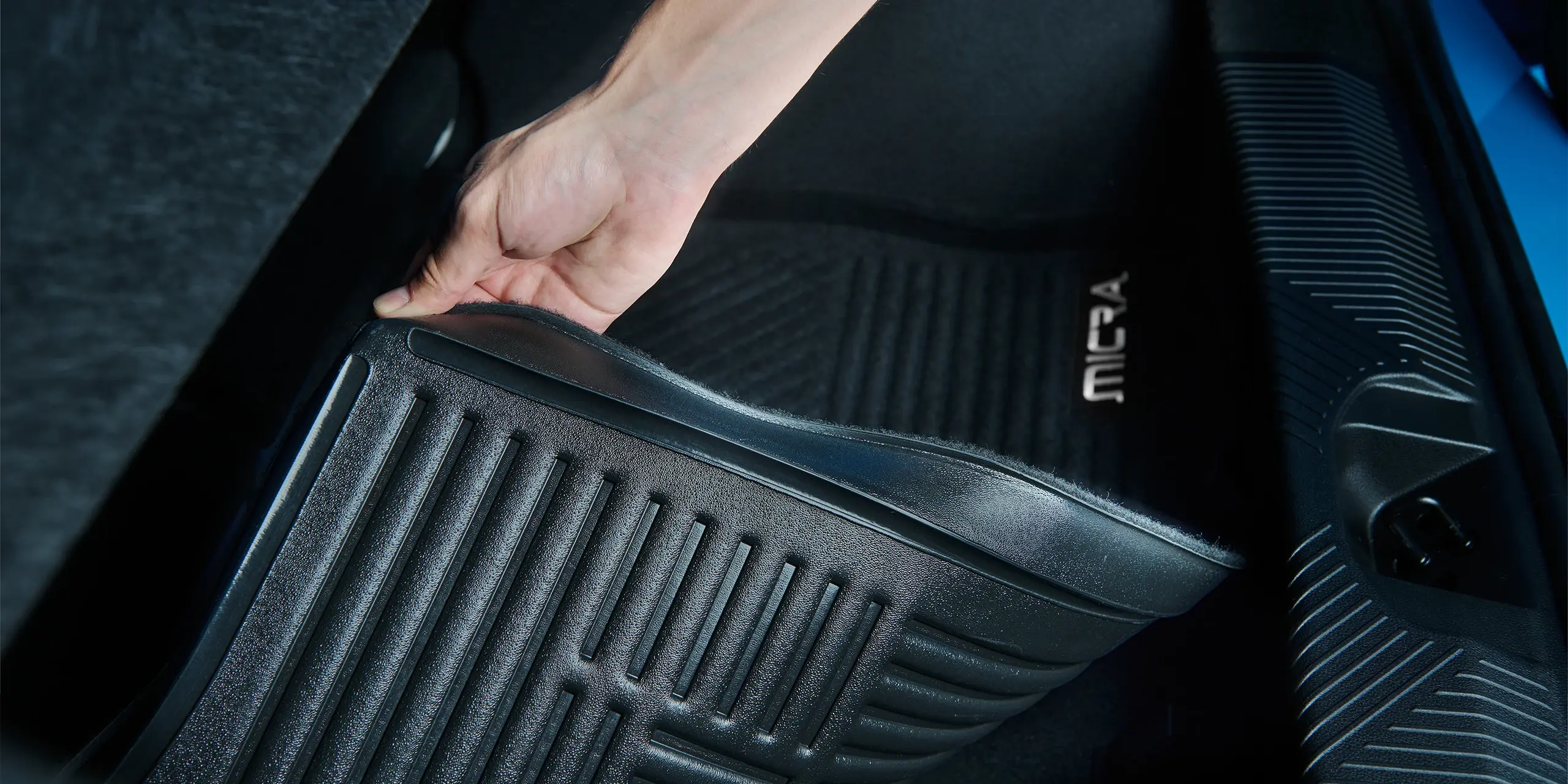 Hand lifting a black rubber car mat with grooves, featuring "MICRA" logo, in a dark car interior.