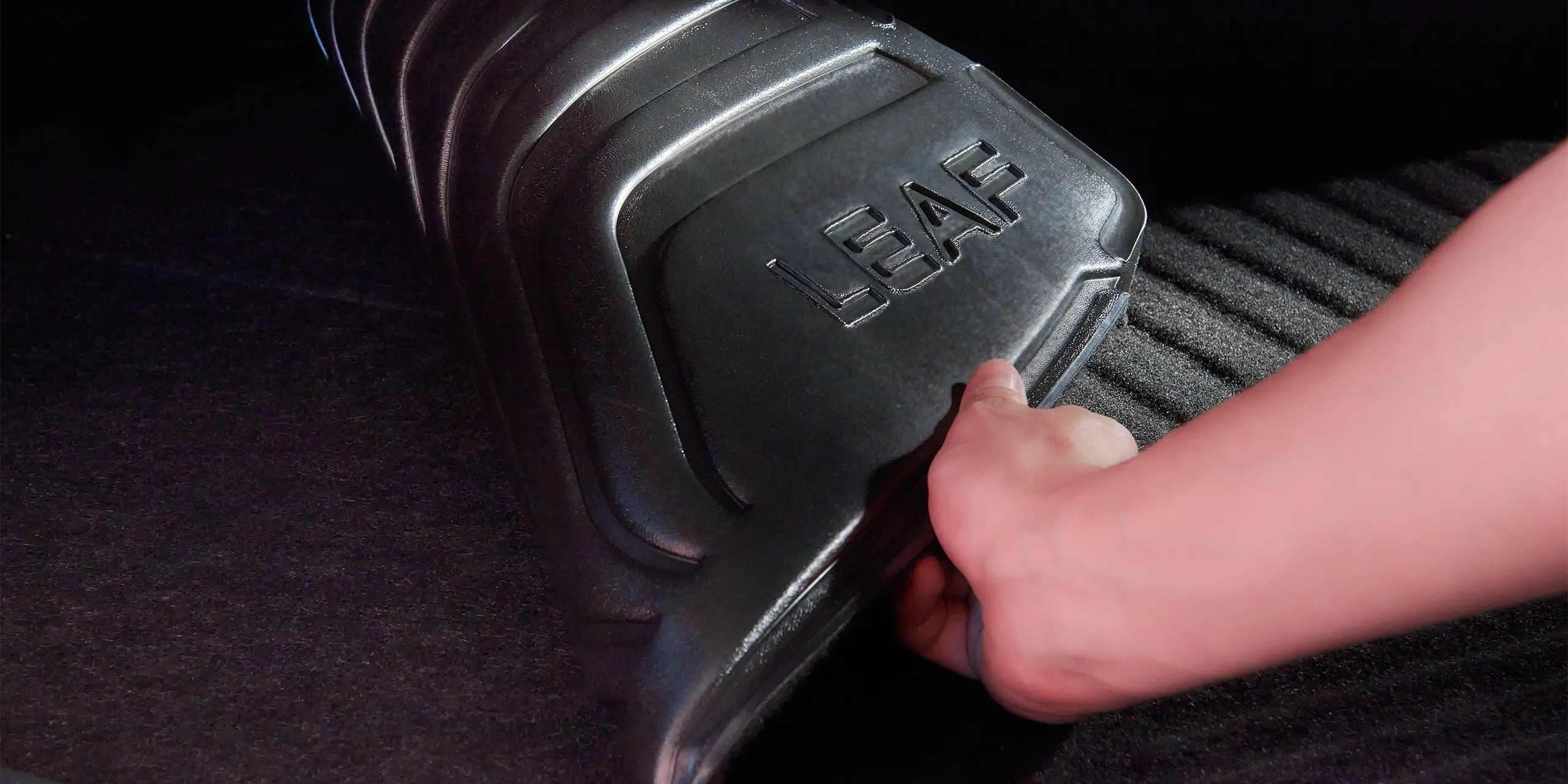 Hand lifting a car floor mat with "LEAF" embossed on it.