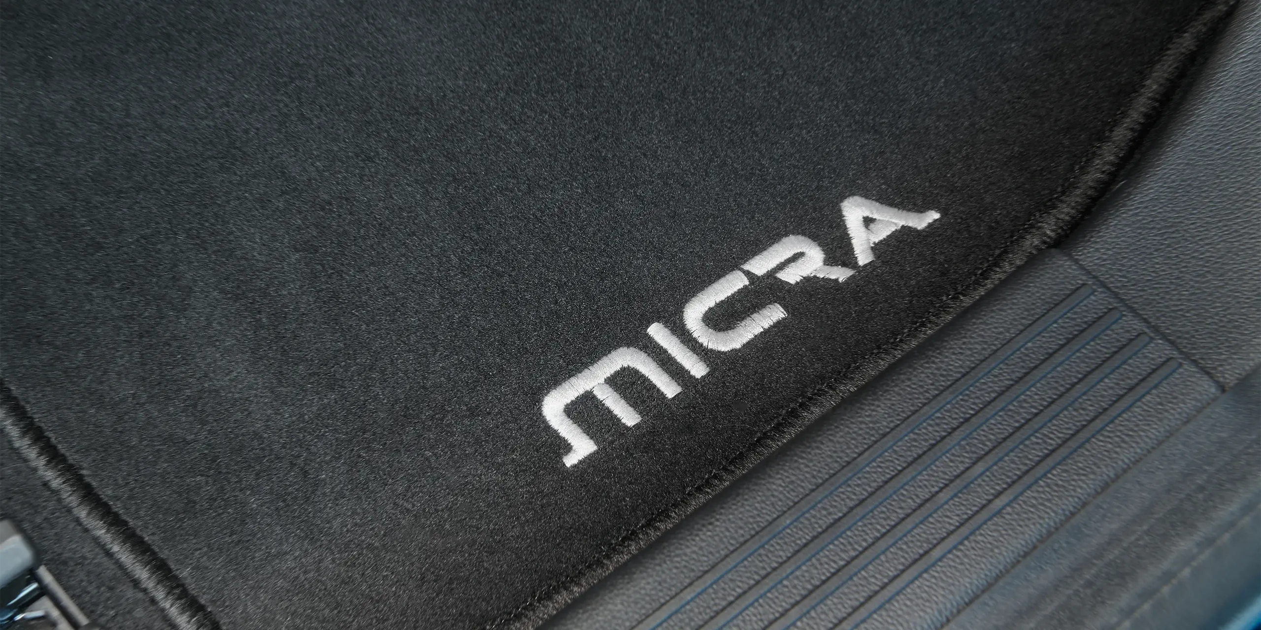 Close-up of a black car mat with "MICRA" embroidered in white, placed on the car floor.